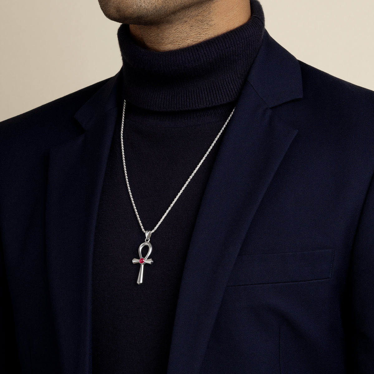 A model wearing a navy blue blazer and turtleneck, modelling a silver ancient Egyptian ankh pendant necklace featuring a round red ruby cabochon stone.